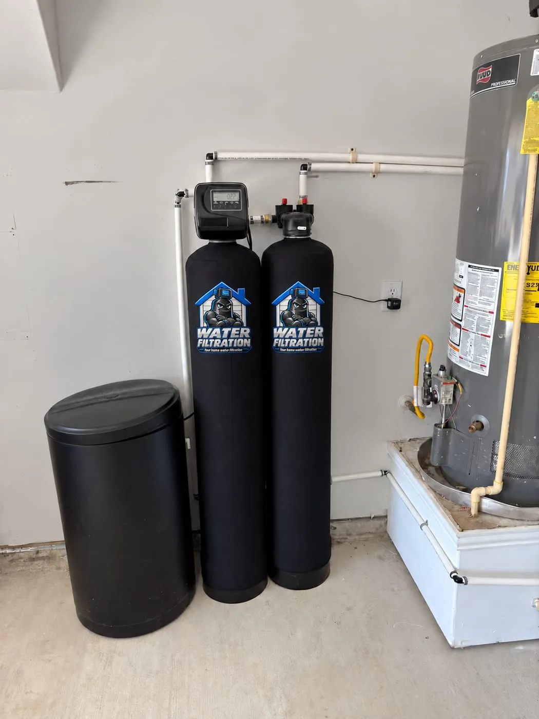Two black filter tanks with Water Filtration logo wraps beside a brine tank and gas water heater in a garage in Bear, DE
