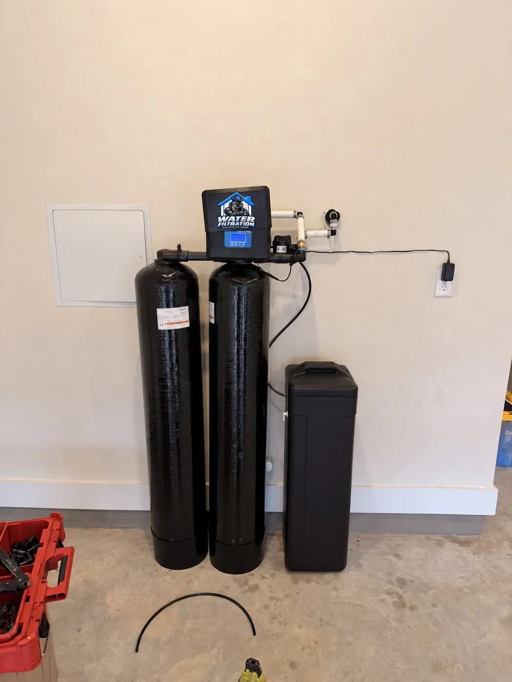 Two filter tanks with a Water Filtration logo valve head and a brine tank installed on a cream garage wall in Dover, DE